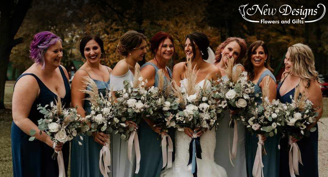 A bride and her bridesmaids stand in a row, the bridesmaids in various shades of dark and soft muted blue, holding bohemian style bouquets with wild greenery, pampas grass and a mix of white flowers such as anemones and roses